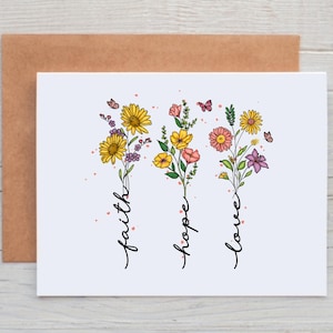 May include: A white greeting card with the words "faith, hope, love" in cursive, each paired with a floral bouquet. The bouquets feature yellow, pink, and purple flowers with butterflies, set against a brown envelope.
