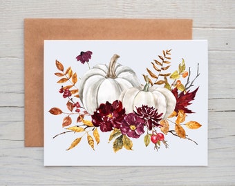 May include: A rectangular greeting card featuring a watercolor illustration of two white pumpkins surrounded by deep burgundy flowers, autumn leaves in shades of orange and brown, and small red berries, accompanied by a plain brown paper envelope.