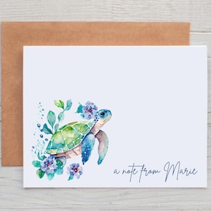 May include: A white stationery card with a watercolor illustration of a sea turtle, surrounded by blue and green foliage and flowers. The text "a note from Marie" is written in a cursive font. A brown envelope is behind the card.