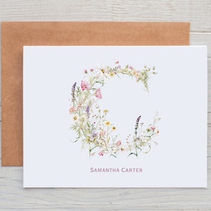 Pu&ograve; includere: Un biglietto bianco con un disegno floreale a forma della lettera C. I fiori sono rosa, bianchi e viola. Il nome "Samantha Carter" &egrave; stampato in rosa nella parte inferiore del biglietto.
