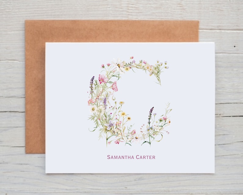 Pu&ograve; includere: Un biglietto bianco con un disegno floreale a forma della lettera C. I fiori sono rosa, bianchi e viola. Il nome "Samantha Carter" &egrave; stampato in rosa nella parte inferiore del biglietto.
