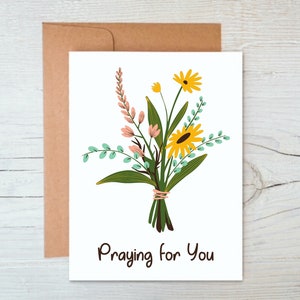 May include: A white card with a bouquet of yellow and pink flowers and green leaves. The text "Praying for You" is printed in black at the bottom of the card.