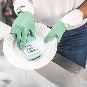 May include: A person wearing green gloves washes a white plate with a green and blue sponge that says "Pop. Fizz. Clean."