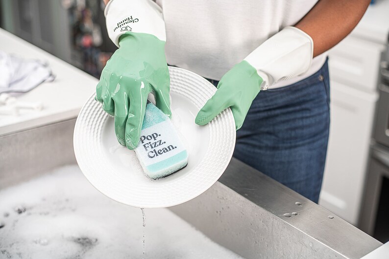 May include: A person wearing green gloves washes a white plate with a green and blue sponge that says "Pop. Fizz. Clean."