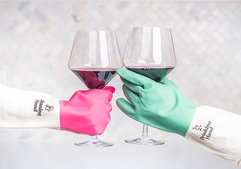 May include: Two hands wearing cleaning gloves, one pink and one green, are holding up glasses of red wine. The gloves have the text "Cleaning Hand" and "Drinking Hand" printed on them.
