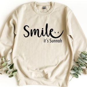 May include: A beige crewneck sweatshirt featuring the black text Smile it's Sunnah, with the letter e extending into a curved line forming a smiling face. The garment has long sleeves, a ribbed collar, and ribbed cuffs, presented in a clean layout.