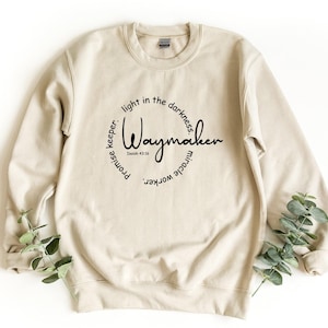 May include: A beige crewneck sweatshirt featuring black text arranged in a circular design. The center reads Waymaker with Isaiah 43:16 below it. The surrounding text includes light in the darkness, promise keeper, and miracle worker.