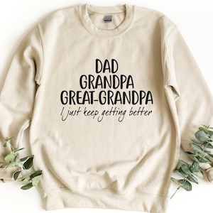 May include: A beige crewneck sweatshirt featuring black text that reads DAD GRANDPA GREAT-GRANDPA I just keep getting better. The garment has long sleeves, a ribbed collar, and ribbed cuffs, presented in a clean, straightforward design.