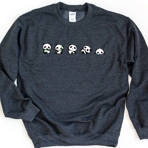 K&ouml;nnte beinhalten: Ein dunkelgrauer Pullover mit einer Reihe von f&uuml;nf kleinen Pandab&auml;ren, die auf der Vorderseite aufgedruckt sind. Die Pandas sind alle nach vorne gerichtet und haben schwarze Augen und Ohren. Die Pandas haben verschiedene Gesichtsausdr&uuml;cke, einige l&auml;cheln, einige sind ver&auml;rgert und einige halten Bambus.