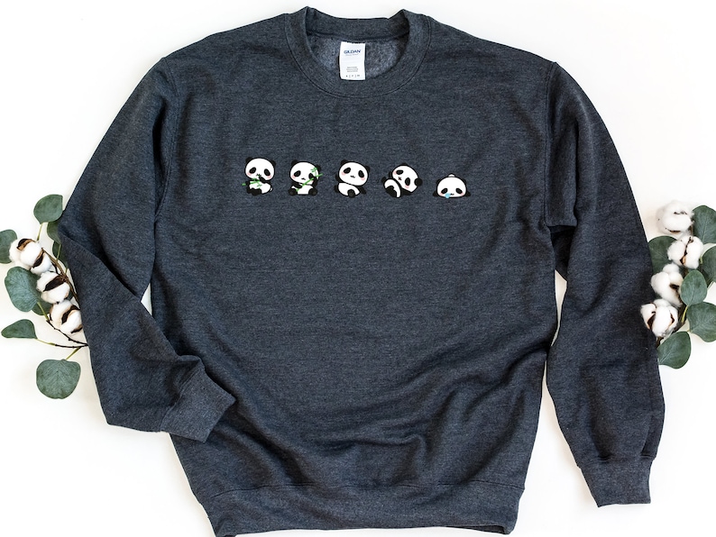 K&ouml;nnte beinhalten: Ein dunkelgrauer Pullover mit einer Reihe von f&uuml;nf kleinen Pandab&auml;ren, die auf der Vorderseite aufgedruckt sind. Die Pandas sind alle nach vorne gerichtet und haben schwarze Augen und Ohren. Die Pandas haben verschiedene Gesichtsausdr&uuml;cke, einige l&auml;cheln, einige sind ver&auml;rgert und einige halten Bambus.