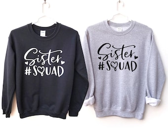 May include: A black sweatshirt with white text reading Sister #SQUAD and a grey sweatshirt with black text reading Sister #SQUAD. Both crewneck garments feature long sleeves, ribbed cuffs, and a heart graphic integrated into the word SQUAD.