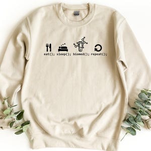 May include: A beige crewneck sweatshirt featuring a black graphic print of icons representing a fork and knife, a bed, a test tube with a molecular structure, and a circular arrow, accompanied by the text eat(); sleep(); biomed(); repeat();.