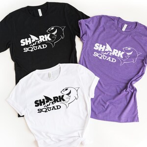 Op de afbeelding: Drie T-shirts met korte mouwen en een ronde hals in zwart, wit en paars. Elk shirt heeft een grafische print van een haai en de tekst SHARK SQUAD. De zwarte en paarse shirts hebben witte tekst en afbeeldingen, het witte shirt heeft zwarte tekst.