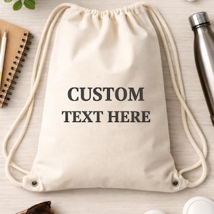 May include: A beige drawstring bag with the text "CUSTOM TEXT HERE" in dark gray. The bag is on a light wood surface, surrounded by accessories like sunglasses, a notebook, and white sneakers.