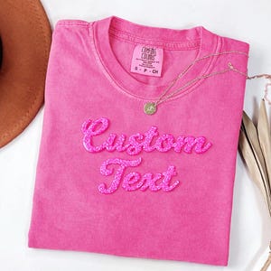 A pink t-shirt with the text "Custom Text" embroidered on it, along with a necklace and a brown hat.
