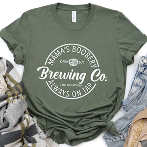 May include: An olive green short-sleeve t-shirt featuring a white circular graphic with the text "MAMA'S BOOBERY BREWING CO. ALWAYS ON TAP" and "OPEN 24/7" surrounding a small barrel icon. The shirt has a crew neckline and rolled sleeves.