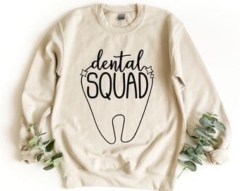 Może przedstawiać: Bluza z okrągłym dekoltem w kolorze piaskowym z czarnym nadrukiem graficznym przedstawiającym zarys zęba oraz napis "dental SQUAD" stylizowaną czcionką. Wzór zawiera małe akcenty w kształcie gwiazdek po obu stronach słowa dental.