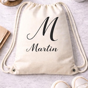 May include: A cream-colored drawstring backpack with the name "Martin" and the letter "M" printed in black. The bag has white drawstrings and metal eyelets. A cup of coffee, a small succulent, and a pair of white sneakers are also visible.