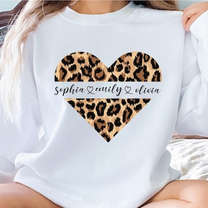 May include: A white long-sleeve sweatshirt featuring a large heart graphic with a brown and black leopard print pattern. A horizontal white stripe across the center of the heart displays the names Sophia, Emily, and Olivia in black script.