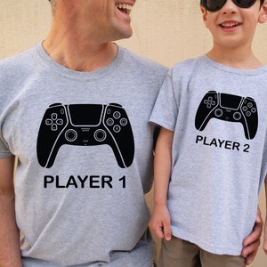 May include: Two grey t-shirts with a black video game controller graphic. The larger shirt says "Player 1" and the smaller shirt says "Player 2".
