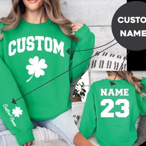 Könnte beinhalten: Ein hellgrünes Langarm-Sweatshirt mit dem weißen Schriftzug CUSTOM und einem vierblättrigen Kleeblatt auf der Vorderseite. Der Ärmel zeigt ein kleines Kleeblatt und einen Namen. Auf der Rückseite steht der weiße Text NAME über der Zahl 23.