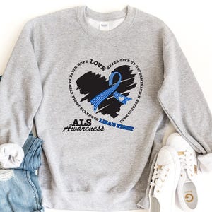 May include: A light gray crewneck sweatshirt featuring a black heart-shaped brushstroke graphic with a blue awareness ribbon. The design includes the text "ALS Awareness" and words like "Faith," "Hope," "Love," "Strength," and "Lira's Fight."