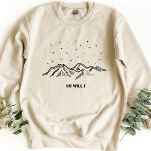 May include: A sand-colored crewneck sweatshirt featuring a black line-art graphic of a mountain range under a starry night sky. The text "SO WILL I" is printed in black capital letters below the mountain illustration on the front of the garment.