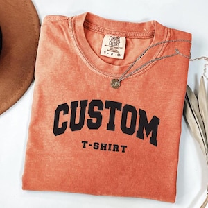 a red t-shirt with the word "CUSTOM" printed on it, along with the text "T-SHIRT" below it. The t-shirt is folded and placed on a white surface, with a brown hat and some leaves visible in the background.
