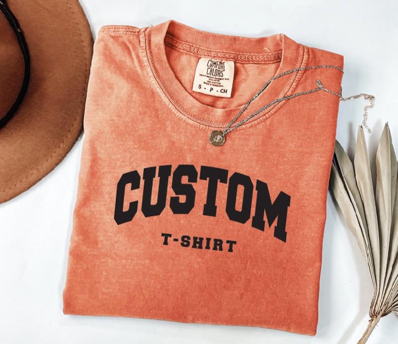 a red t-shirt with the word "CUSTOM" printed on it, along with the text "T-SHIRT" below it. The t-shirt is folded and placed on a white surface, with a brown hat and some leaves visible in the background.