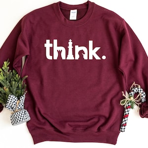 Può includere: Una felpa bordeaux a maniche lunghe con girocollo che presenta la scritta bianca "think.", in cui le lettere "h" e "k" sono stilizzate come pezzi degli scacchi, nello specifico una torre, un re e un cavallo, con un punto alla fine.