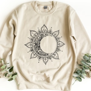 Puede incluir: Una sudadera beige con un diseño de mandala negro que presenta una luna creciente. El mandala es intrincado y detallado, con un patrón circular de hojas y flores.
