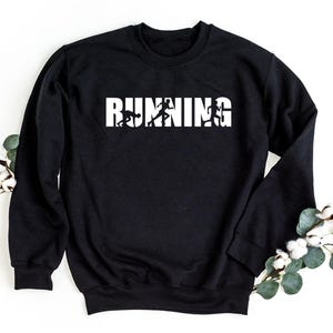 Op de afbeelding: Een zwart sweatshirt met lange mouwen en een ronde hals met de witte tekst RUNNING op de borst. Het ontwerp bevat drie zwarte silhouetten van een persoon in verschillende fasen van een hardloopbeweging, verwerkt in de witte letters.