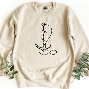 May include: A sand-colored crewneck sweatshirt featuring a black graphic design of an anchor intertwined with the cursive word hope. The anchor includes a small heart detail at its base, and the design is centered on the front of the garment.