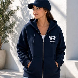 Può includere: Una felpa con cappuccio e zip blu navy, dotata di cappuccio con coulisse, maniche lunghe, polsini a coste e due tasche anteriori. Il testo CUSTOM TEXT LOGO è ricamato in bianco sul petto sinistro. È indossato anche un berretto da baseball blu navy.