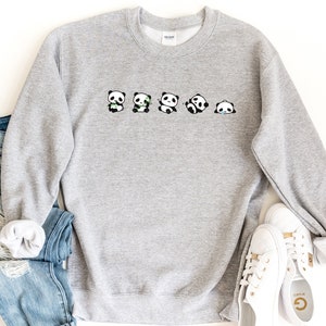 K&ouml;nnte beinhalten: Ein grauer Pullover mit einer Reihe von f&uuml;nf schwarzen und wei&szlig;en Pandab&auml;ren, die verschiedene Gesichtsausdr&uuml;cke zeigen. Die Pandas halten Bambusstangen in ihren Pfoten.