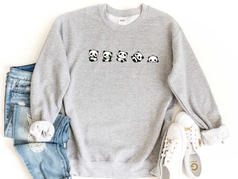 K&ouml;nnte beinhalten: Ein grauer Pullover mit einer Reihe von f&uuml;nf schwarzen und wei&szlig;en Pandab&auml;ren, die verschiedene Gesichtsausdr&uuml;cke zeigen. Die Pandas halten Bambusstangen in ihren Pfoten.
