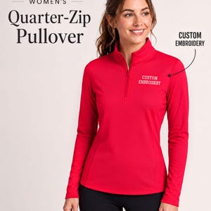 May include: A bright red long-sleeve quarter-zip pullover featuring a high collar and a zipper closure. The text "CUSTOM EMBROIDERY" is stitched in white thread on the upper left chest area, accompanied by a black arrow pointing to the text.