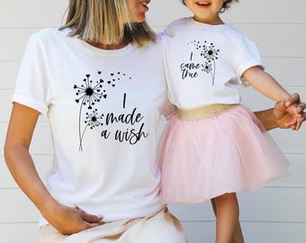 May include: Two white short-sleeved t-shirts featuring black dandelion graphics with heart-shaped seeds. The larger shirt displays the text I made a wish, while the smaller shirt displays the text I came true, paired with a light pink tulle skirt.