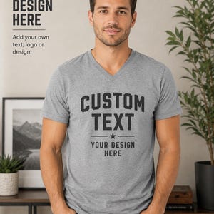 Op de afbeelding: Een grijs gemêleerd T-shirt met korte mouwen en V-hals, voorzien van de zwarte tekst CUSTOM TEXT, een ster en horizontale lijnen boven de woorden YOUR DESIGN HERE. Links bovenaan staat de tekst YOUR DESIGN HERE Add your own text, logo or design!