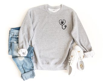 May include: A light grey crewneck sweatshirt featuring a black graphic on the left chest. The graphic depicts a stethoscope shaped into a heart with a paw print in the center. The sweatshirt is paired with light blue distressed denim jeans and white sneakers.