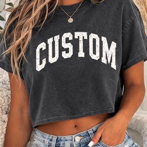 Op de afbeelding: Een antracietgrijs kort T-shirt met het woord CUSTOM in een verweerd wit lettertype in universiteitsstijl op de borst. Het kledingstuk heeft een ronde hals, korte mouwen en een zoom met rafelige rand die boven de taille valt.