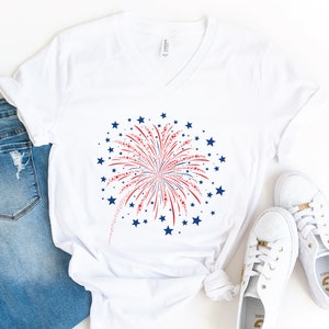 Può includere: Maglietta bianca a collo a V con un motivo di fuochi d'artificio rosso, bianco e blu. I fuochi d'artificio sono circondati da stelle blu.