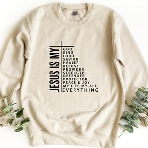 Op de afbeelding: Een beige sweater met ronde hals en zwarte tekst in de vorm van een kruis. De tekst luidt: JESUS IS MY GOD, KING, LORD, SAVIOR, HEALER, REFUGE, PROVIDER, STRENGTH, DEFENDER, PROTECTOR, PEACE & JOY, MY LIFE, MY ALL, EVERYTHING.