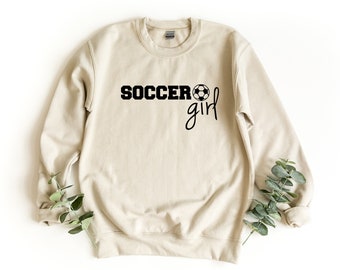 Może przedstawiać: Beżowa bluza z czarnym napisem "Soccer girl" i czarnym nadrukiem piłki nożnej.