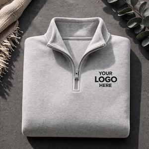 Könnte beinhalten: Ein gefalteter hellgrauer Sweatshirt mit einem Kragen mit Viertelreißverschluss. Der Text "YOUR LOGO HERE" ist in Schwarz auf der Vorderseite aufgedruckt. Das Sweatshirt besteht aus einem weichen Material und ist ordentlich gefaltet. Eine neutralfarbene Decke und Grünzeug befinden sich im Hintergrund.
