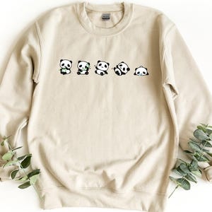 May include: A sand-colored crewneck sweatshirt featuring a graphic print of five small black and white pandas in various poses, including sitting, eating bamboo, and rolling, arranged in a horizontal line across the chest area of the garment.