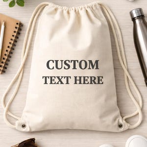 May include: A cream-colored canvas drawstring backpack featuring the black text CUSTOM TEXT HERE printed in the center. The bag has thick, matching cream-colored rope straps that cinch the top opening and are secured with metal grommets.