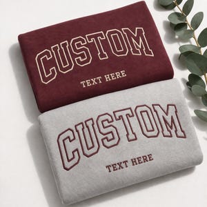 Two t-shirts with the word "CUSTOM" printed on them, along with the text "Text Here" below it. The t-shirts are in different colors - maroon and gray.