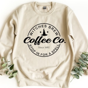 Può includere: Una felpa beige con una grafica circolare nera che dice "Witches Brew Coffee Co. Since 1692 Stop in for a Spell" con un cappello da strega e stelle all'interno del cerchio.