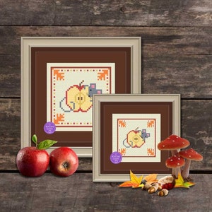 May include: Two framed cross-stitch patterns featuring a mouse and an apple. The smaller frame is in front of the larger frame. Both frames have a brown background and a cream-colored mat. The smaller frame is on a wooden surface with fall-themed decorations, including acorns, leaves, and mushrooms.